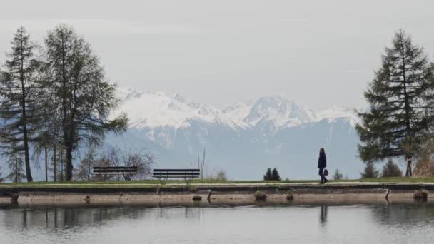 Женщина ходит по набережной со скамейкой рядом с озером Этанг Лонг в Кранс-Монтане, Швейцария в солнечный весенний день. Великолепные снежные Альпы на заднем плане. Общий вид — стоковое видео