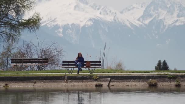 Atraktivní dívka sedí na dřevěné lavici s úžasným výhledem na jezero a hory. Žena, která si užívá malebného místa. Rekreace a relaxace — Stock video