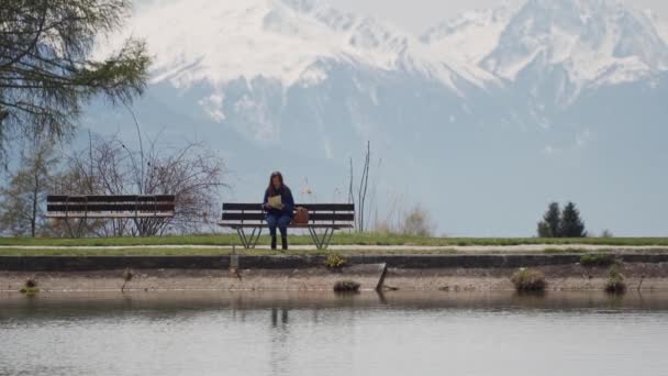 Tourist Girl zit op een bankje bij een bergmeer, neemt een kaart uit een rugzak en begint te kijken naar de kaart — Stockvideo