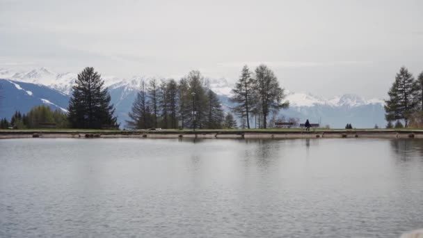 Girl gets up from a bench on the shore of a picturesque lake and leaves. Pine trees and mountains are in the background — Stock Video