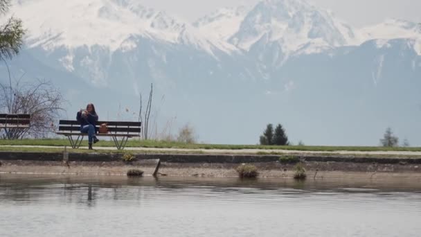 Mujer joven se sienta en un banco de madera y toma fotos por teléfono inteligente increíble vista sobre el lago de montaña — Vídeos de Stock