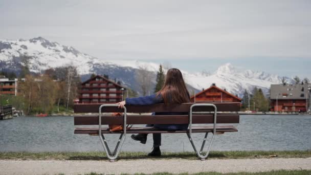 Ung kvinna sitter på en träbänk med fantastisk utsikt över sjön och bergen och avslappnad. Kvinna njuter av en pittoresk plats. Bakifrån — Stockvideo