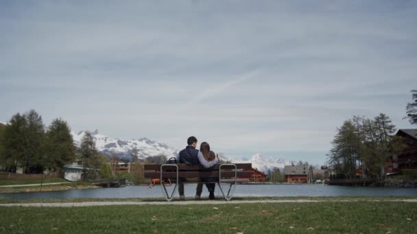 Giovane coppia coccole sul lungolago panca in legno con bella vista sulle montagne. Vista posteriore. Piano generale — Video Stock