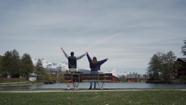 Pareja feliz enamorada de pie junto con las manos levantadas en el banco de madera junto al lago con hermosas vistas a la montaña. Vista trasera. Plan general — Vídeos de Stock