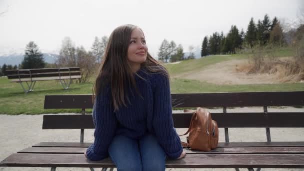 La joven sonriente se sienta en un banco de madera con una vista increíble sobre el lago y las montañas y se relajó en el soleado día de primavera. Mujer disfrutando de un lugar pintoresco . — Vídeos de Stock