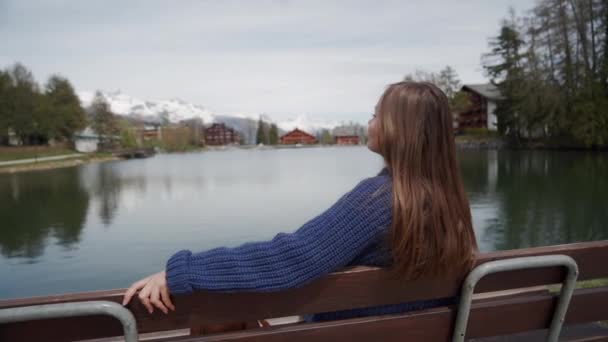 Mujer joven se sienta en un banco de madera con una vista increíble sobre el lago y las montañas y relajado en el soleado día de primavera. Mujer disfrutando de un lugar pintoresco. Vista trasera — Vídeos de Stock