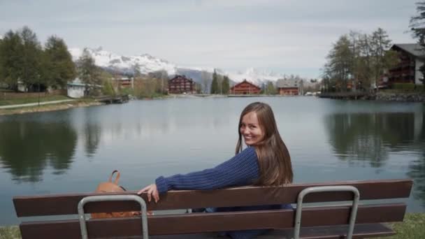 Jovem mulher sentada no banco de madeira à beira do lago com vista panorâmica para a montanha. Então menina olhando para trás e sorrindo lindamente para a câmera — Vídeo de Stock