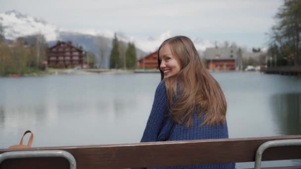 Hermosa mujer de pelo largo sentada en un banco de madera junto al lago con vistas panorámicas a la montaña. Entonces chica mirando hacia atrás y sonriendo maravillosamente a la cámara — Vídeos de Stock