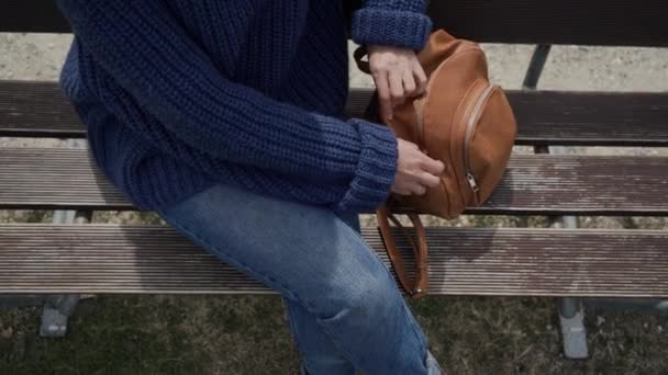 Tourist girl sits on a bench by a mountain lake, takes out a map from brown backpack and starts to look at map. Close up — Stock Video