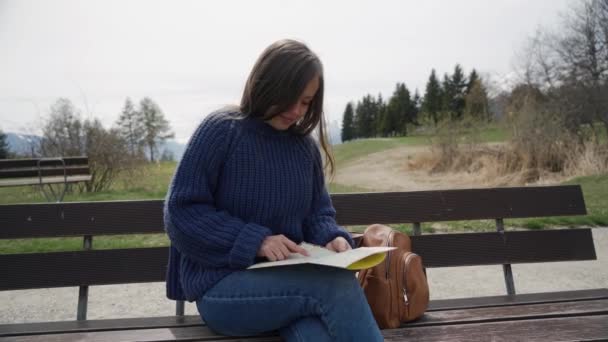 Žena seděla na dřevěné lavici s mapou papíru. Turisté hledají místo určení na mapě papíru, ukazovali na místa a porozhlédli se kolem. Zavři to. Pohled zepředu — Stock video