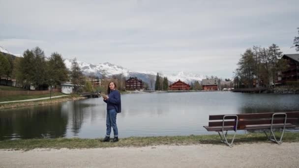 Kvinna i casual kläder stående med papper karta i händerna på Lakeside med naturskön utsikt över bergen. Girl söker plats med hjälp av karta och titta runt. Allmän plan — Stockvideo