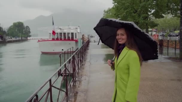 Счастливая женщина в жёлтом пальто с чёрным зонтиком в горошек идёт по дождю вдоль живописного озера Анси, Франция. Вид сзади — стоковое видео