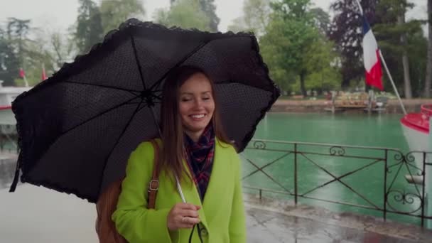 Beautiful girl in a yellow coat stands in the rain, twists a cute black umbrella, smiles and flirts. Lake Annecy, France. Portrait — ストック動画