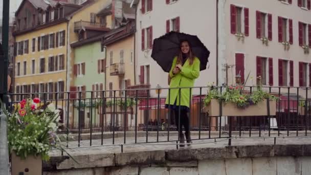 Yağmurlu bir gün. Sarı paltolu ve şemsiyeli genç bir kadın köprüde duruyor ve gülümsüyor. Annecy. Fransa — Stok video