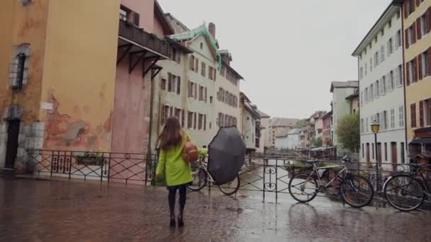 Menina feliz bonita andando na ponte perto dos edifícios antigos, brincando com seu guarda-chuva preto. Dia chuvoso. Morena jovem mulher vestindo casaco amarelo e vestido — Vídeo de Stock
