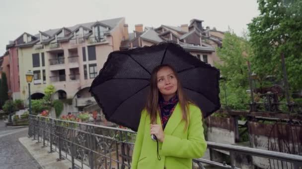 Die hübsche Frau steht bei Regen unter einem schwarzen Regenschirm auf der Brücke. Porträt — Stockvideo