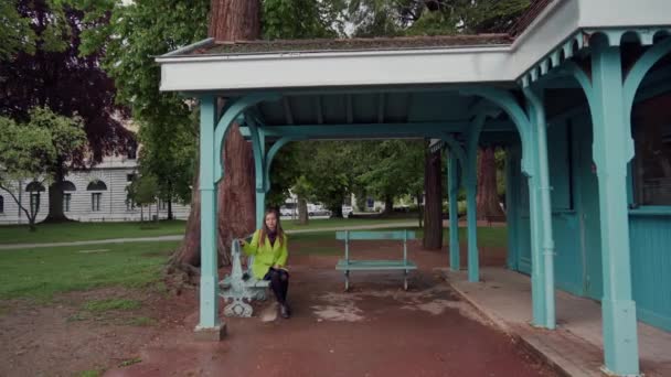 Krásná dívka ve žlutém kabátě sedí na lavičce pod střechou modrého kovového pavilonu v deštivém dni — Stock video