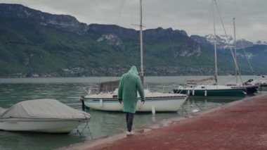 Man in light green raincoat walking near sailboats moored on lake in the mountains in foggy weather. White swan swimming in a pond