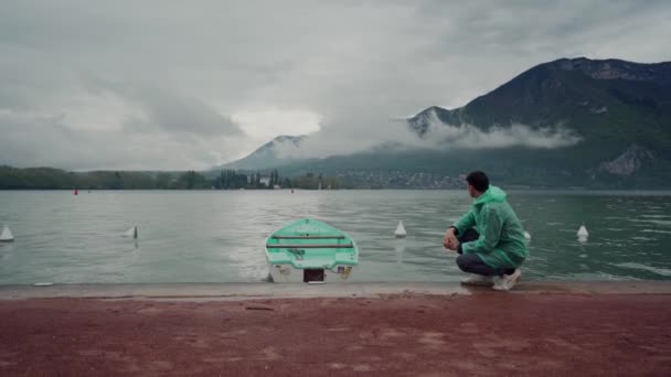 Egy férfi egy mentolos esőkabátban ül egy tavon a hegyekben, egy hasonló színű kikötött hajó mellett. Esős nap. Annecy-tó, Franciaország — Stock videók