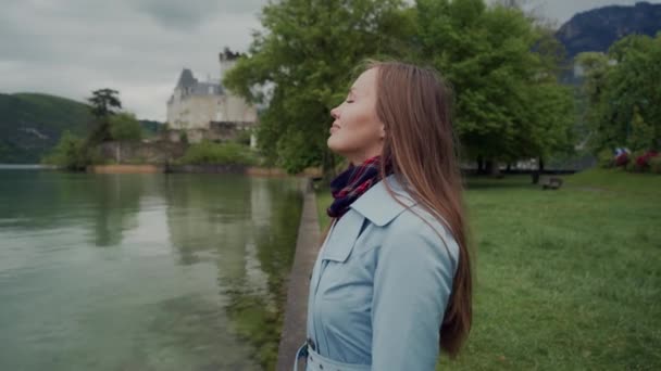 Närbild av attraktiv flicka står i profil på stranden av sjön Annecy. Hon bär blå rock och ser framåt med leende. Castle Ruphy är på bakgrunden. Duingt, Frankrike — Stockvideo