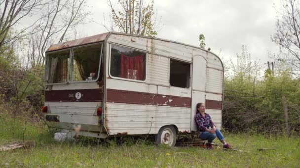 Mujer joven en ropa casual meditativamente se sienta cerca de la vieja caravana abandonada — Vídeo de stock