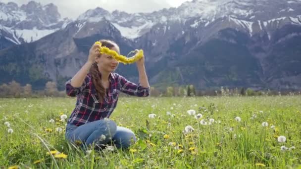 Cerca de hermosa joven teje una corona de flores de diente de león prado y se prueba en sí misma en el fondo de las montañas — Vídeo de stock