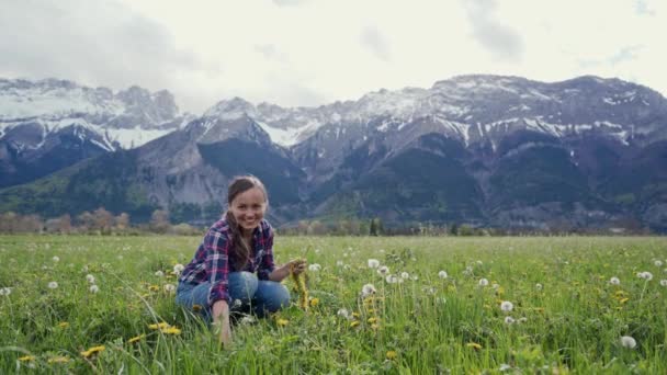 Menina feliz tece uma coroa de flores de dente-de-leão amarelo sentado no prado no fundo das montanhas. Tradições nacionais eslavas — Vídeo de Stock