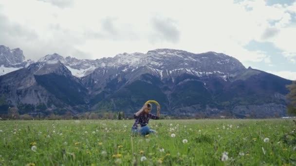 Jonge vrouw probeert paardenbloem krans op zichzelf op bergen achtergrond. Algemeen plan. Landschappelijk berglandschap — Stockvideo