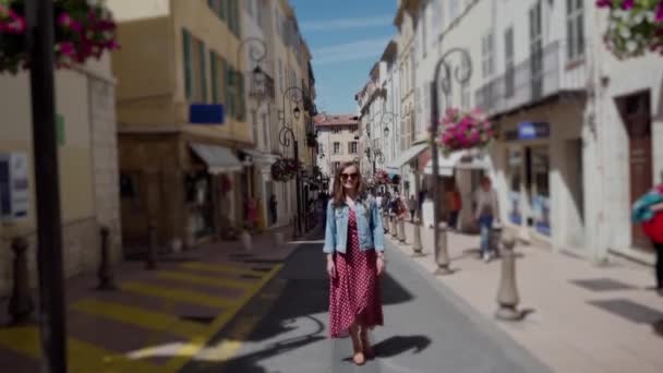 Mulher bonita nova com óculos de sol andando no meio da rua velha da cidade em um dia ensolarado. Viajante feminina vestindo vestido romântico vermelho polka-dot. Visão de perspectiva borrada da rua estreita com — Vídeo de Stock