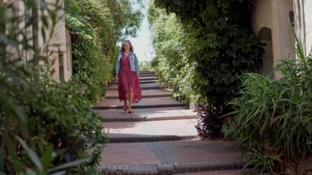 Женщина в солнцезащитных очках спускается по каменной лестнице в маленьком французском городе Антиб. Девушка ходит и одета в романтическое красное платье в горошек и джинсовую куртку. Счастливая модель в хорошем настроении на отдыхе — стоковое видео