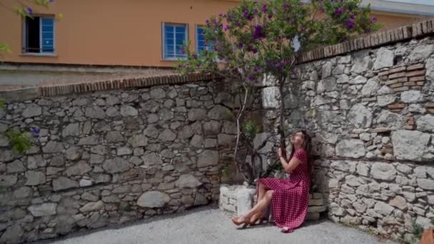 Mujer bonita joven en vestido rojo sentado en la cama de flores de piedra bajo el árbol violeta floreciente en el soleado día de primavera en la vieja ciudad antigua — Vídeos de Stock