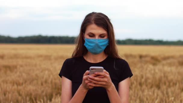 漂亮的年轻女子戴着医疗面罩 站在田野里看着电话 保护自己不受感染 — 图库视频影像