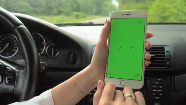 女性の手は車のハンドルに手で緑の画面で携帯電話を保持します 女の子のドライバーは彼女の手にクロマキーで携帯電話を保持ホイールに座っています — ストック動画