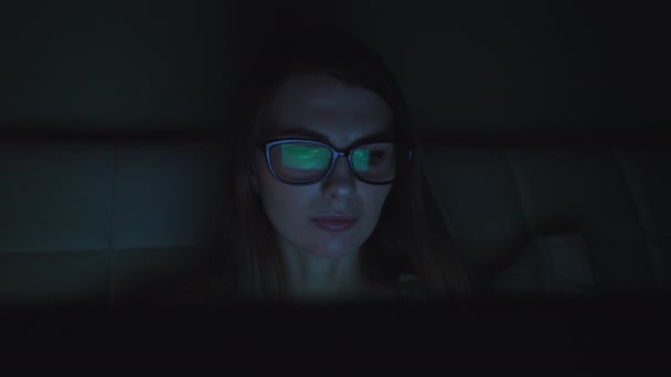 Portret młodej dziewczyny w okularach, dziewczyna leżąca na kanapie z laptopem i wpisująca wiadomość. W nocy. Poduszka pod głowę dziewczyny. Nie ma światła w salonie. Światło ekranu na twarzy dziewczyn — Wideo stockowe
