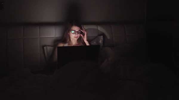 Mulher de óculos trabalha em um laptop no escuro. retrato de uma menina passar o tempo no computador tarde da noite — Fotografia de Stock