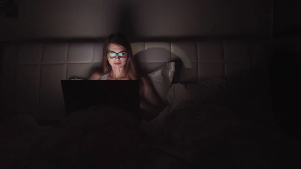 Mulher de óculos trabalha em um laptop no escuro. retrato de uma menina passar o tempo no computador tarde da noite — Fotografia de Stock