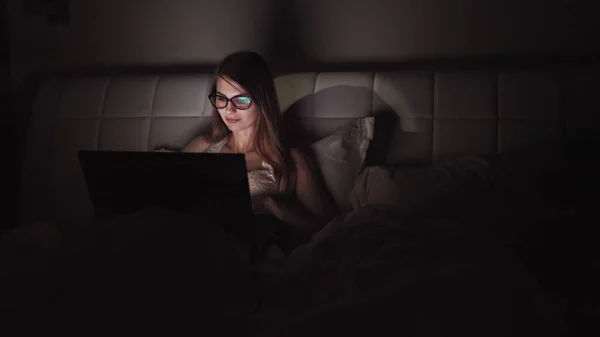 Mulher de óculos trabalha em um laptop no escuro. retrato de uma menina passar o tempo no computador tarde da noite — Fotografia de Stock