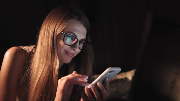 Onnellinen tuhatvuotinen tyttö makaa vatsassa sängyssä hymyillen katsoen älypuhelinta illalla, nuori inkivääri teini tilalla mobiililaite nauraa sosiaalisen median internet-sisältöä pukeutunut viihtyisä pyjama — kuvapankkivalokuva