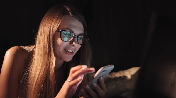 Onnellinen tuhatvuotinen tyttö makaa vatsassa sängyssä hymyillen katsoen älypuhelinta illalla, nuori inkivääri teini tilalla mobiililaite nauraa sosiaalisen median internet-sisältöä pukeutunut viihtyisä pyjama — kuvapankkivalokuva