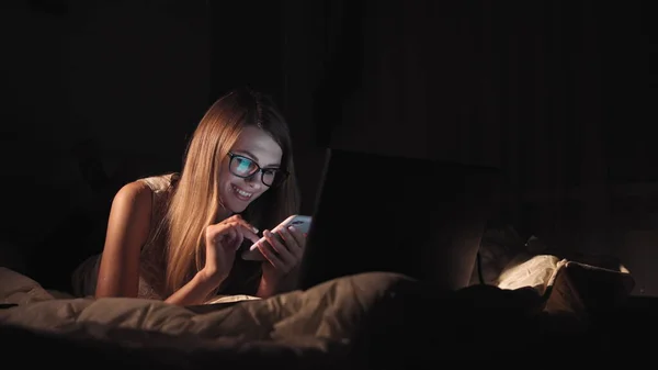 Vista lateral de un adolescente feliz navegando y viendo los medios de comunicación en un teléfono inteligente acostado en una cama en la noche en casa — Foto de Stock