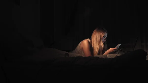 Chica feliz seleccionando y viendo vídeo en un teléfono inteligente acostado en la cama en la noche en casa — Foto de Stock