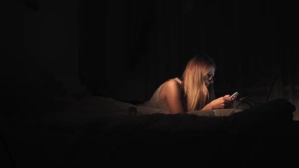 Chica feliz seleccionando y viendo vídeo en un teléfono inteligente acostado en la cama en la noche en casa — Foto de Stock