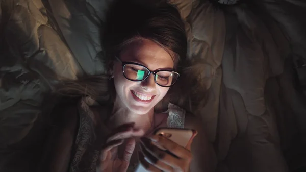 Girl watching comedy movie at night in bed. Tablet smart phone or laptop glowing on womans face. Pretty teen laughing ad smiling at film at sleep over in bed room