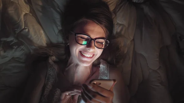 Chica viendo películas de comedia por la noche en la cama. Tableta de teléfono inteligente o portátil que brilla en la cara de las mujeres. bastante adolescente riendo anuncio sonriendo en película en dormir encima en cama habitación — Foto de Stock
