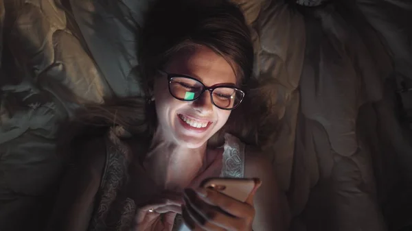 Ragazza che guarda film comici di notte a letto. Tablet smart phone o laptop incandescente sul viso delle donne. Bella teen ridere annuncio sorridente a film a dormire sopra in camera da letto — Foto Stock