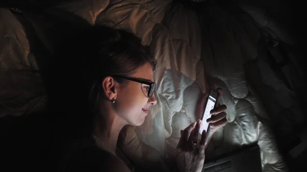 Tenåringsjente på berøringsskjerm Smart Laptop og hodetelefoner på sengen, student i mørkt rom med mobilt nattlys, en ung kvinne som leser og leser om kvelden. stockbilde