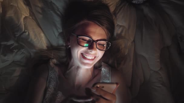 Dziewczyna ogląda komedię w nocy w łóżku. Tablet smartfon lub laptop świecące na twarzy kobiet. Ładny nastolatek śmieje się reklama uśmiechając się do filmu w spać nad w łóżku — Wideo stockowe