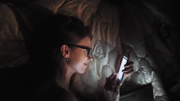 Підліткова дівчинка на сенсорному екрані розумна лаптоп і навушники на ліжку, студент у темній кімнаті з мобільним нічним світлом, молода жінка вчиться і читає ввечері. — стокове відео