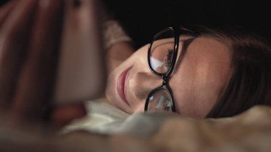 Gözlüklü kadın akıllı telefona bakar ve yatağa uzanır, yüzüne gülümser.