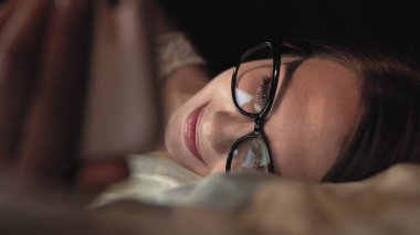 Gözlüklü kadın akıllı telefona bakar ve yatağa uzanır, yüzüne gülümser.